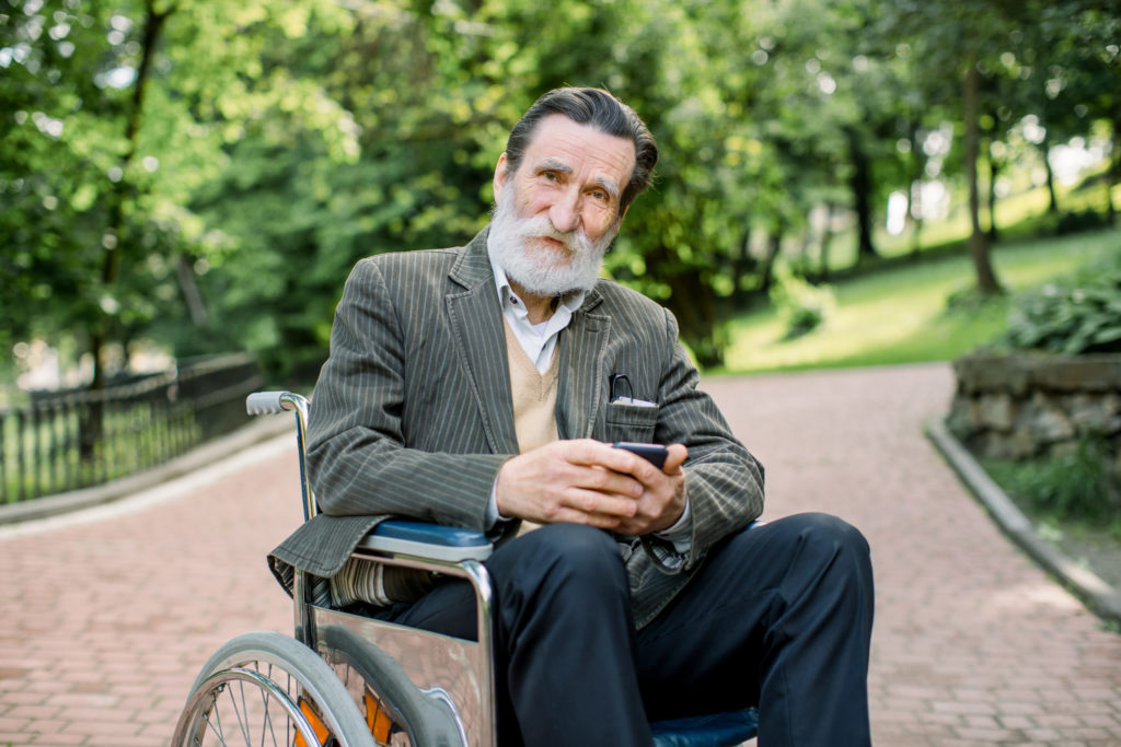 Handicapped elderly bearded man sitting in a wheelchair in city park outdoors, holding his mobile