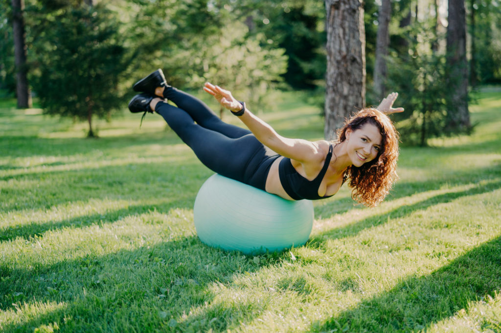 Woman with European appearance balances on fitness ball stretches arms.